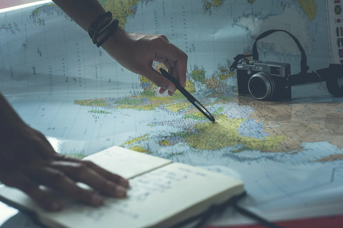 Une main trace un itinéraire au stylo sur une carte du monde, à côté d’un carnet