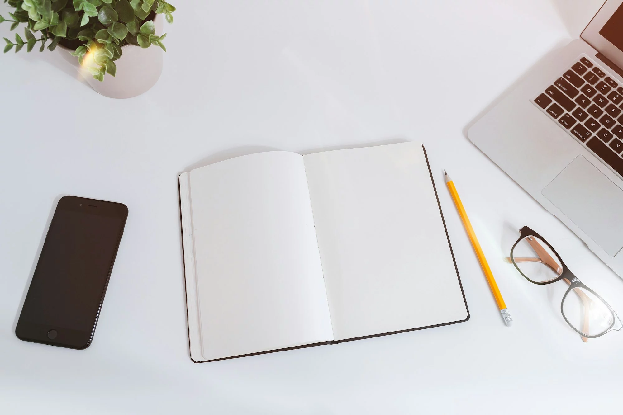 Un carnet ouvert, un crayon et des lunettes posés sur un bureau blanc, à côté d’un ordinateur portable